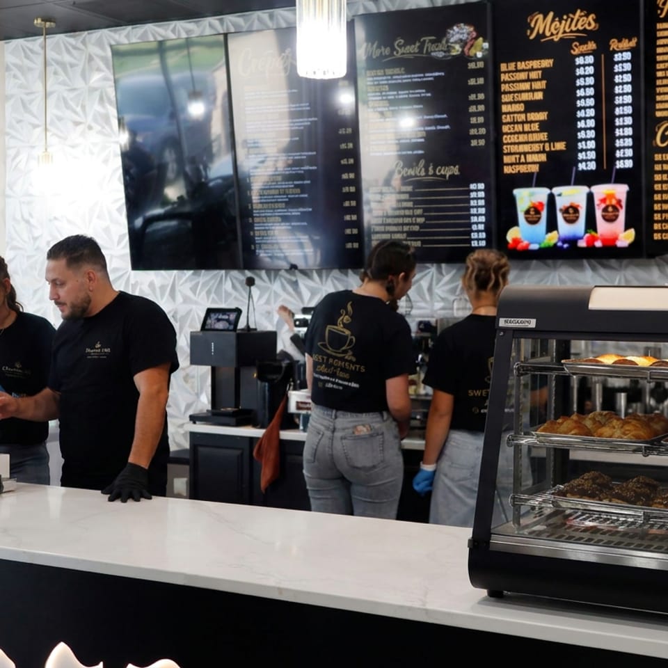 Café counter with staff, pastries in a glass display case, espresso equipment, and menu boards advertising drinks and crepes.