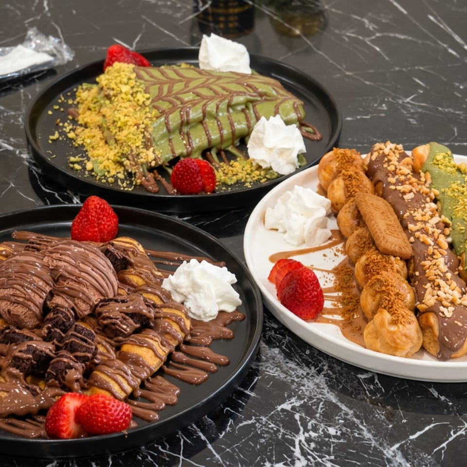 Three dessert plates featuring chocolate and pistachio waffles, churros, strawberries, whipped cream, sauces, and crushed nuts on a marble table.