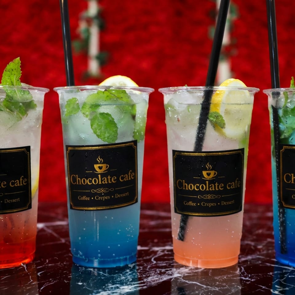 Four colorful iced mint drinks with lemon garnishes and straws, labeled Chocolate cafe, arranged against a festive red background.
