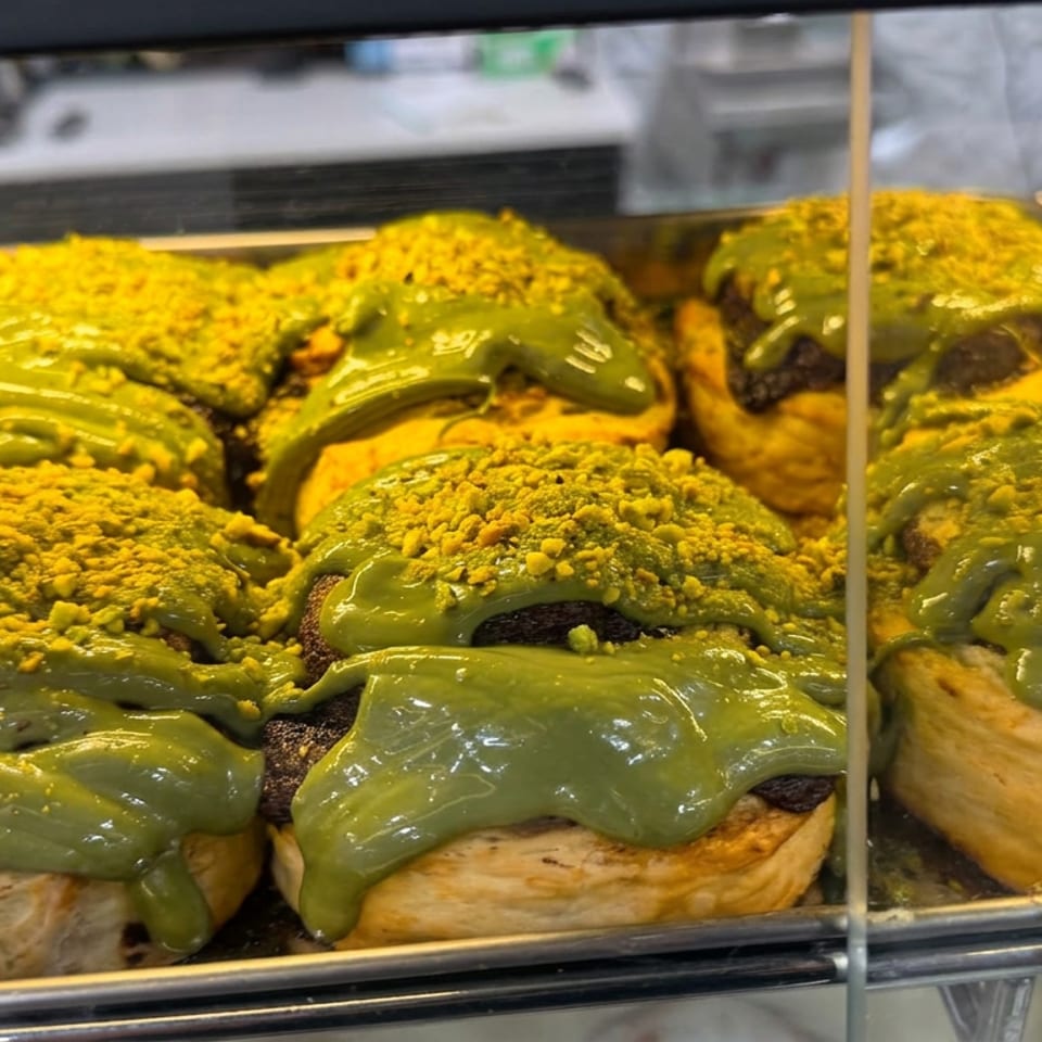 Tray of golden pastries topped with glossy green icing and crushed pistachios behind a bakery display case.