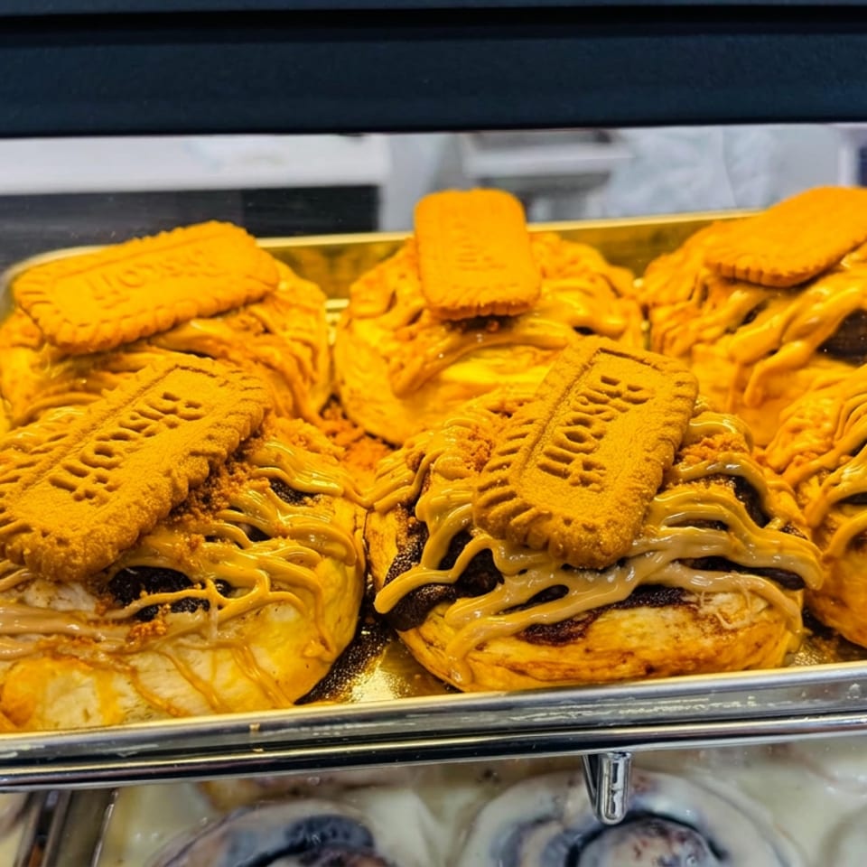 Tray of golden pastries topped with Biscoff biscuits and generous caramel drizzle in a bakery display case.